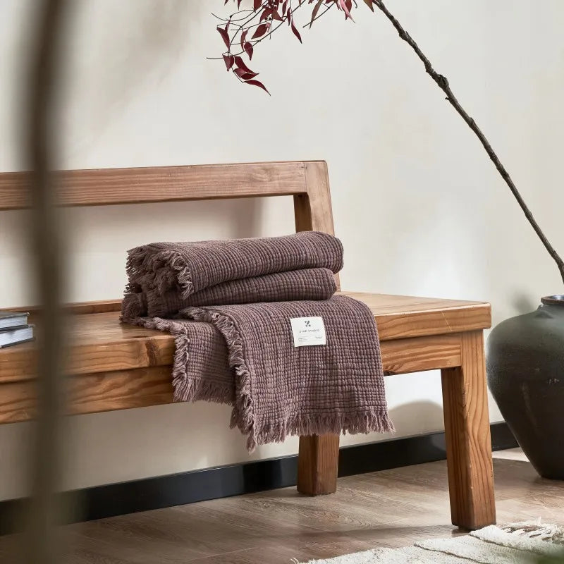 Folded brown blanket on a wooden bench with a vase in the background Maillard cotton gauze blanket with tassels, four-layer muslin throw for sofa, bed, and boho home decor