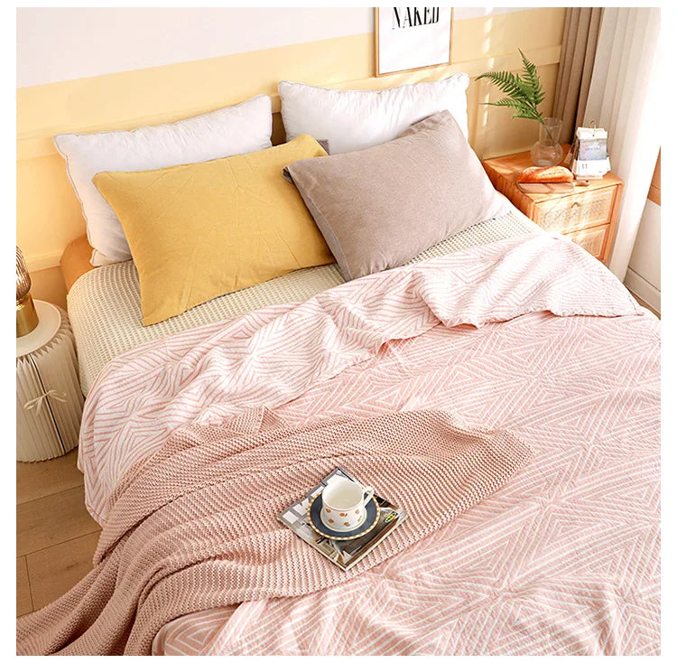 Bed with pink blanket, yellow and gray pillows, and a small table with a cup. Boho Lightweight microfiber summer blanket for bed or sofa, cooling breathable throw for home, dorm, or office naps with geometric jacquard pattern.