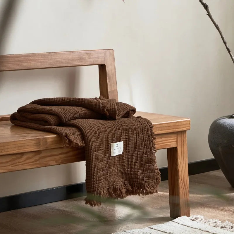 Brown textured blanket on a wooden bench with a neutral background Maillard cotton gauze blanket with tassels, four-layer muslin throw for sofa, bed, and boho home decor