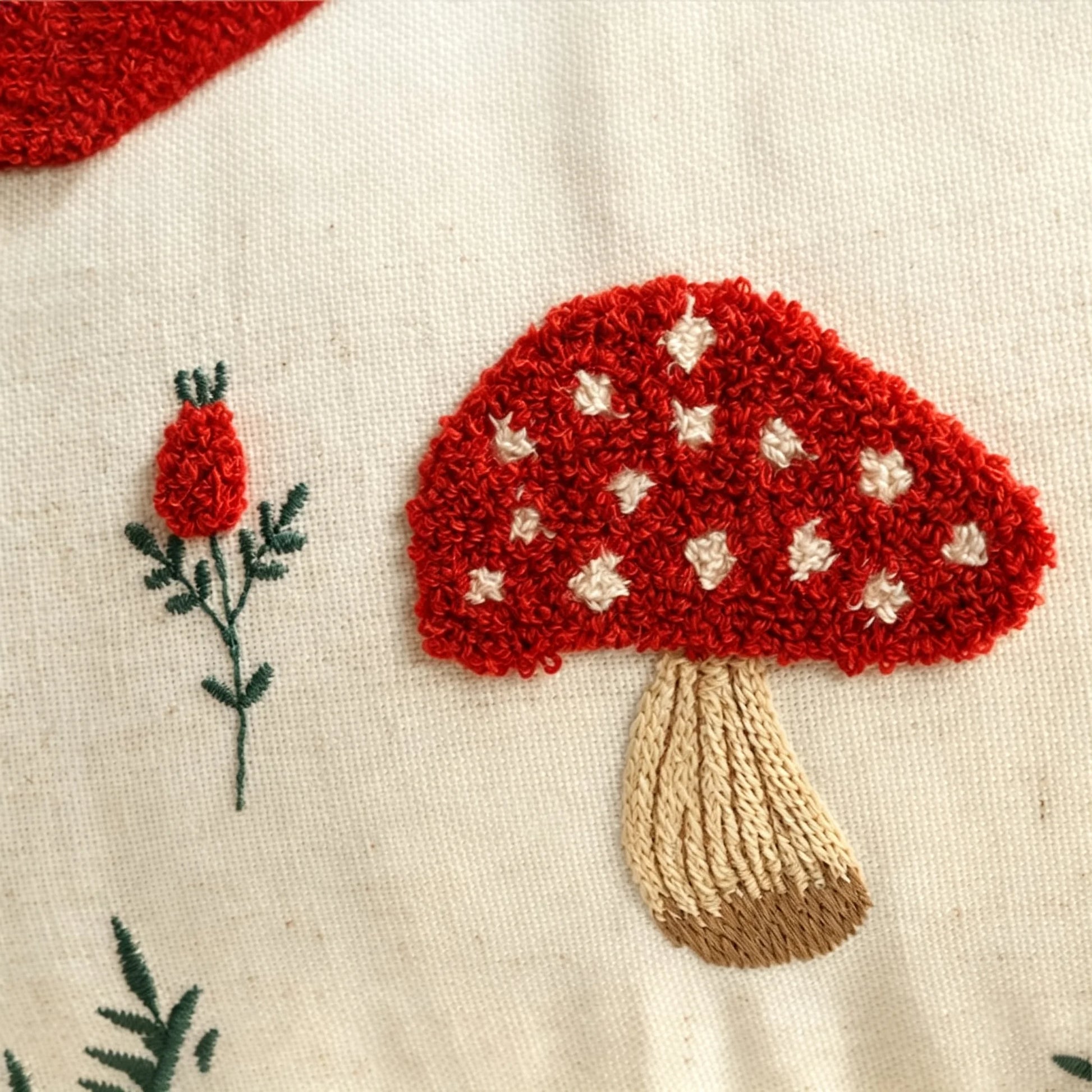 Close-up of a red embroidered mushroom with white spots and a tan stem on a cream fabric background, with a small red flower stitched nearby. Boho fall home decor.
