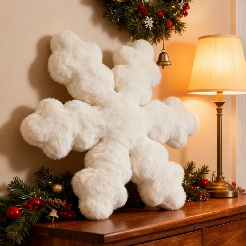 A large fluffy white snowflake-shaped decorative pillow or plush ornament displayed on a wooden console table in a warmly lit holiday room, with a lit table lamp, green garland, red berries, and a hanging wreath in the background.