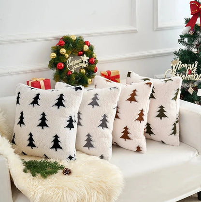 Decorative pillows with Christmas tree patterns on a white couch, with Christmas trees and wreath in the background.