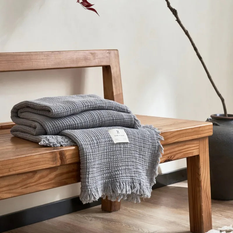 Gray textured blanket on a wooden bench with a neutral background Maillard cotton gauze blanket with tassels, four-layer muslin throw for sofa, bed, and boho home decor