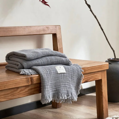 Gray textured blanket on a wooden bench with a neutral background Maillard cotton gauze blanket with tassels, four-layer muslin throw for sofa, bed, and boho home decor