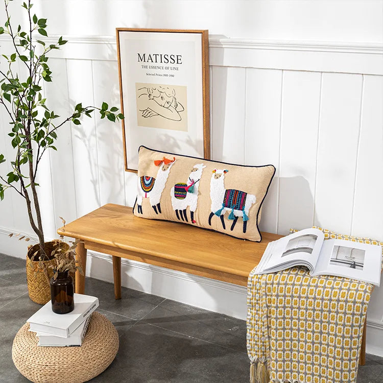 Decorative pillow with llama design on a wooden bench in a room with a plant and framed picture. Bohemian embroidered throw pillow cover with geometric design, cotton-blend cushion for sofa, bed, or modern home decor.