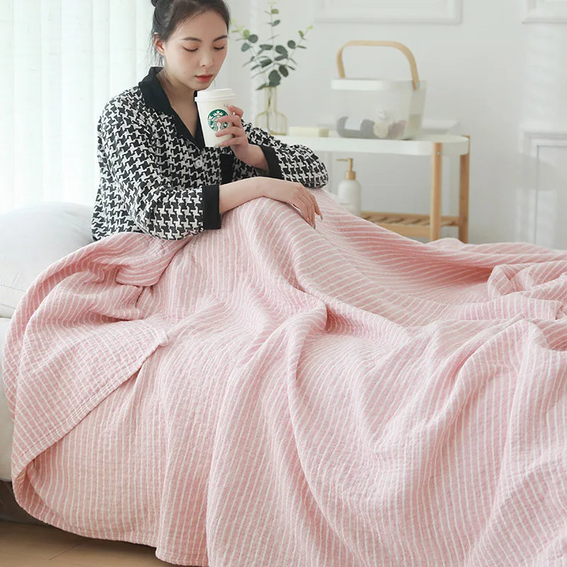 Woman sitting on a couch under a pink blanket, holding a coffee cup. Boho Lightweight microfiber summer blanket for bed or sofa, cooling breathable throw for home, dorm, or office naps with geometric jacquard pattern.