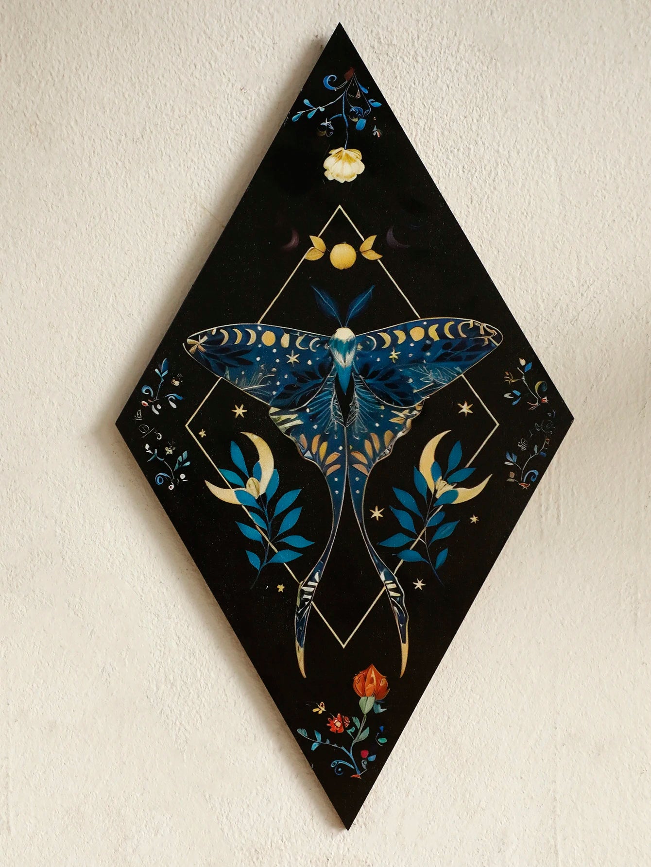 Decorative wall art with a blue moth design on a black diamond-shaped canvas. Rustic colorful wood wall art panel with moon, stars, and butterfly engraving, displayed above a sofa in a boho-styled living room