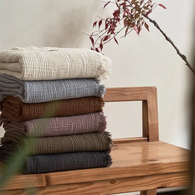 Stack of folded blankets in various colors on a wooden surface with a branch in the background. Maillard cotton gauze blanket with tassels, four-layer muslin throw for sofa, bed, and boho home decor