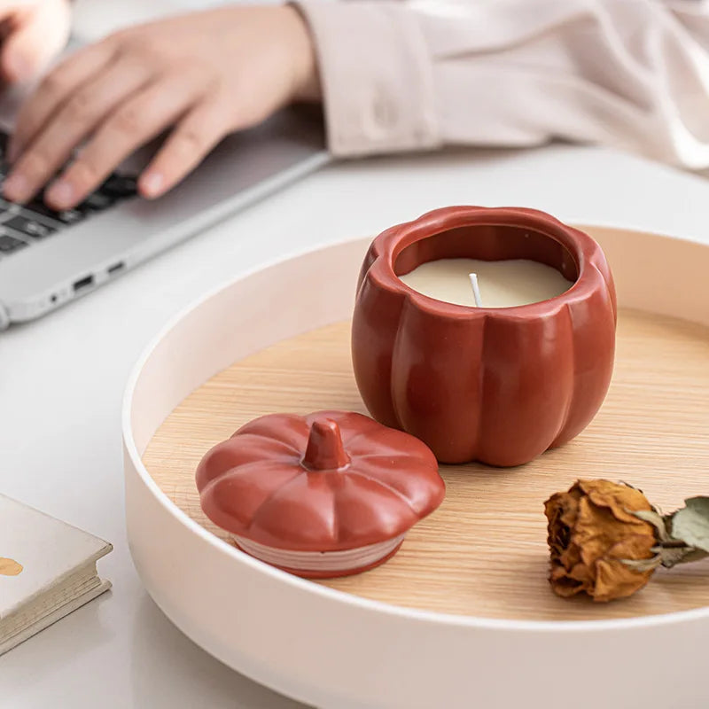 Pumpkin-shaped ceramic candle on a wooden tray with a person using a laptop in the background. fall home decor