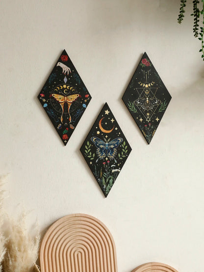 Three diamond-shaped wall art pieces with celestial designs on a white wall. Colorful wooden wall decor featuring moon phases, stars, and butterflies, ideal for boho or gothic home interiors