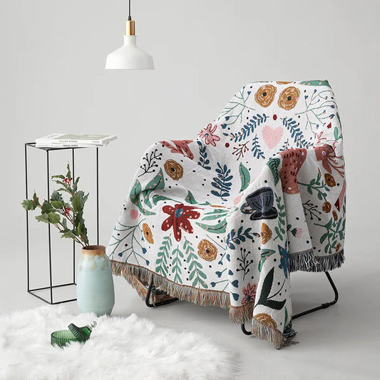 Floral-patterned blanket draped over a chair with a small table and plant in the background. Deer forest jacquard woven blanket with floral and woodland tapestry design – boho throw for sofa or bed, soft cotton blend decorative bedspread for cozy home décor.