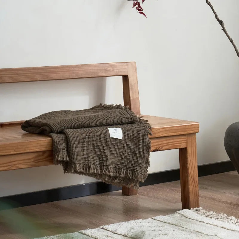 Wooden bench with a brown blanket draped over it against a plain wall. Maillard cotton gauze blanket with tassels, four-layer muslin throw for sofa, bed, and boho home decor