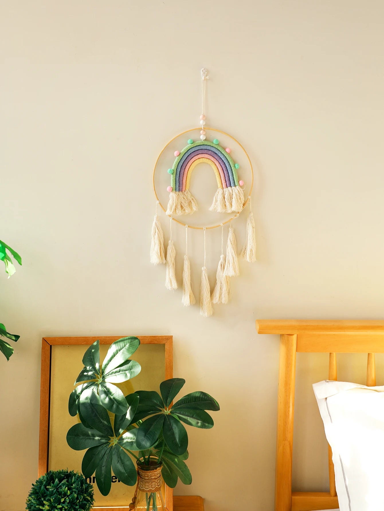 Decorative rainbow dreamcatcher hanging on a wall with a plant and bed frame in the background. Handmade macrame rainbow wall hanging boho home decor