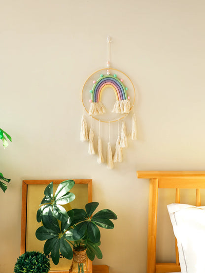 Decorative rainbow dreamcatcher hanging on a wall with a plant and bed frame in the background. Handmade macrame rainbow wall hanging boho home decor
