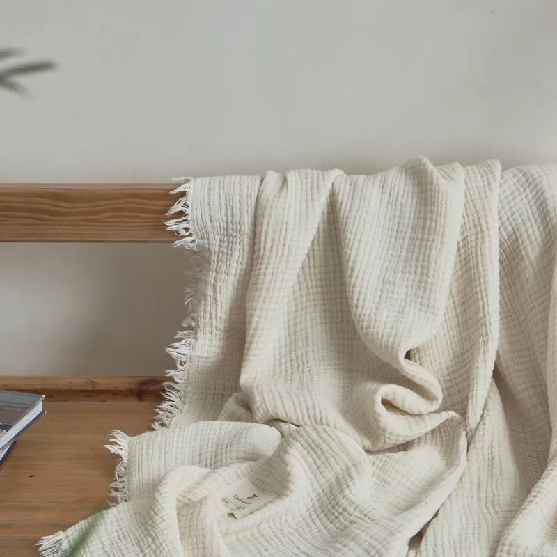 Beige textured blanket draped over a wooden surface with a neutral background Maillard cotton gauze blanket with tassels, four-layer muslin throw for sofa, bed, and boho home decor