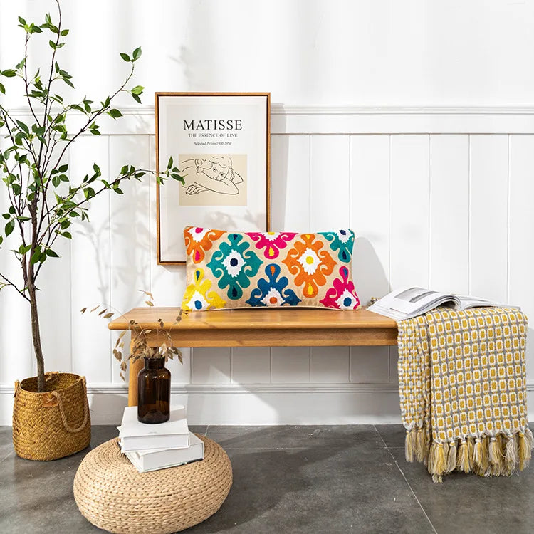 Colorful patterned pillow on a wooden bench with a plant and books in the background. Bohemian embroidered throw pillow cover with geometric design, cotton-blend cushion for sofa, bed, or modern home decor.