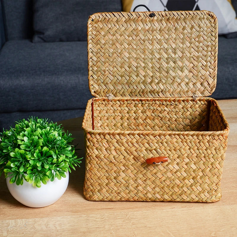 Woven storage basket with a lid on a wooden surface next to a small potted plant. Handmade seagrass rattan storage box with lid, rectangular woven organizer basket for makeup, jewelry, office, and boho home decor