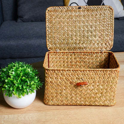 Woven storage basket with a lid on a wooden surface next to a small potted plant. Handmade seagrass rattan storage box with lid, rectangular woven organizer basket for makeup, jewelry, office, and boho home decor