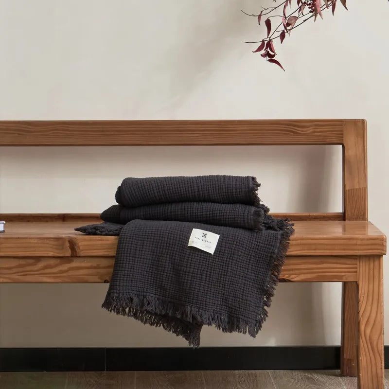 Black textured blanket draped over a wooden bench with a neutral background Maillard cotton gauze blanket with tassels, four-layer muslin throw for sofa, bed, and boho home decor