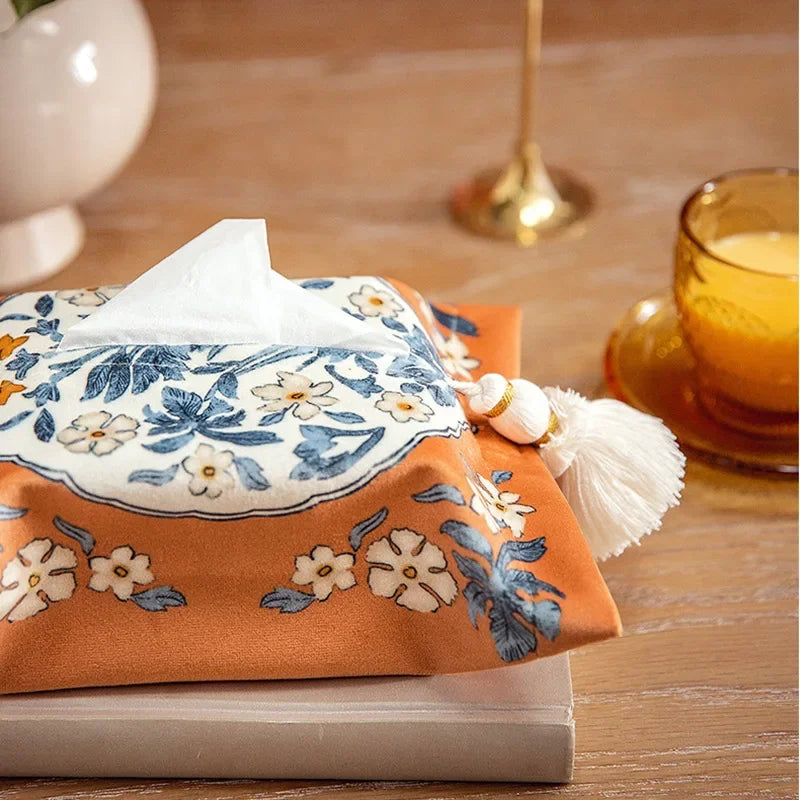 Floral-patterned tissue box cover on a wooden surface with a cup and candle in the background. boho home decor, reusable tissue bag, cotton tassel tissue holder, plush tissue organizer, bathroom tissue box cover, car tissue case, American style tissue pouch, soft fabric tissue holder, decorative tissue box, housewarming gift, eco-friendly storage