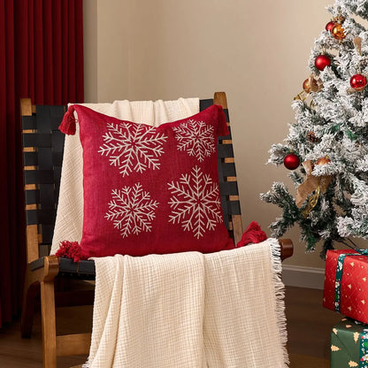 Red pillow with snowflake patterns on a chair next to a decorated Christmas tree. Embroidered Merry Christmas pillow cover with snowflake design – festive red and white holiday cushion for sofa or bedroom décor.
