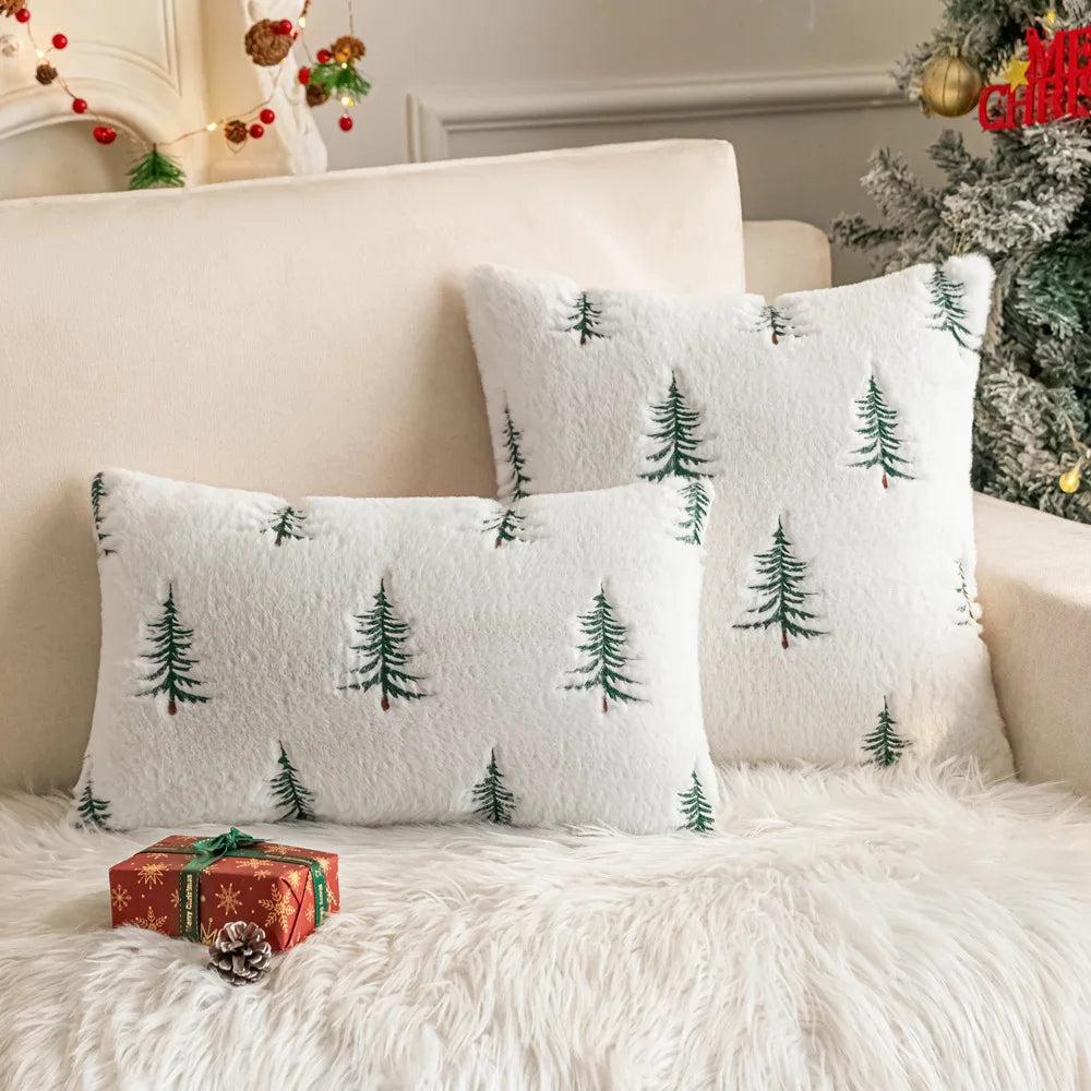White pillows with green tree patterns on a white couch with a Christmas tree in the background.