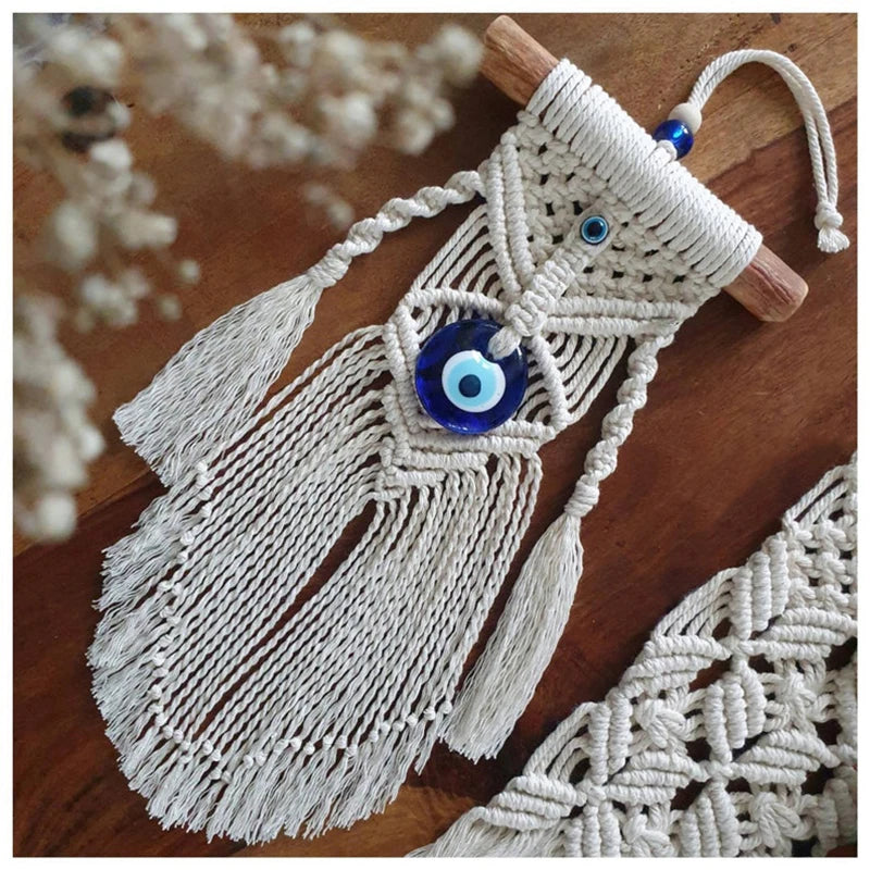 A white macrame wall hanging with blue evil eye beads and tassels, displayed on a wooden surface with dried flowers in the background.
