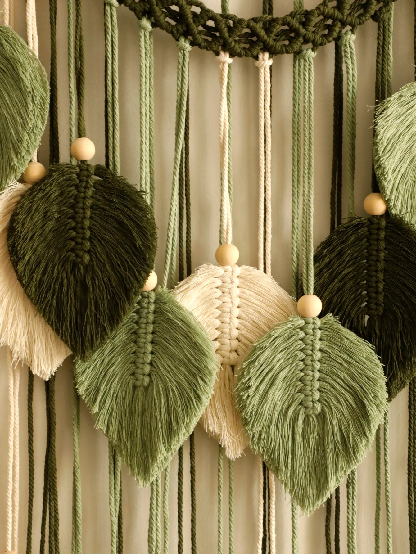 Macrame wall hanging with green, beige, and brown yarn and wooden beads on a striped background.