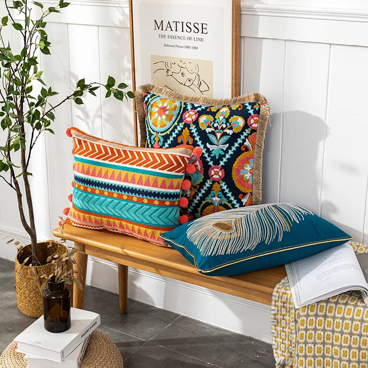 Colorful pillows on a wooden bench with a plant and books in the background.Bohemian embroidered throw pillow cover with geometric design, cotton-blend cushion for sofa, bed, or modern home decor.