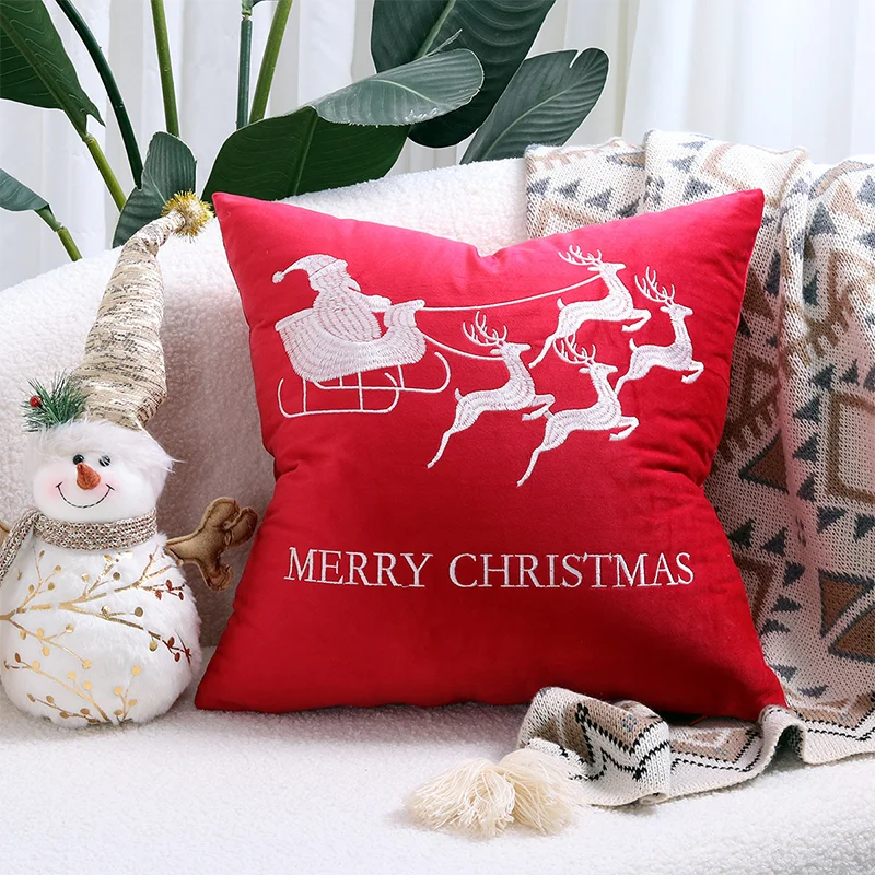 Red Christmas pillow with reindeer and sleigh design on a couch with festive decorations. Embroidered Merry Christmas pillow cover with snowflake design – festive red and white holiday cushion for sofa or bedroom décor.