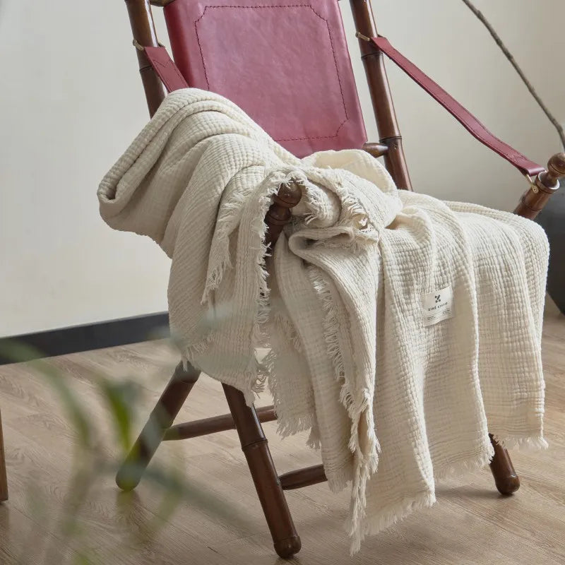 White textured blanket draped over a wooden chair with a pink cushion. Maillard cotton gauze blanket with tassels, four-layer muslin throw for sofa, bed, and boho home decor