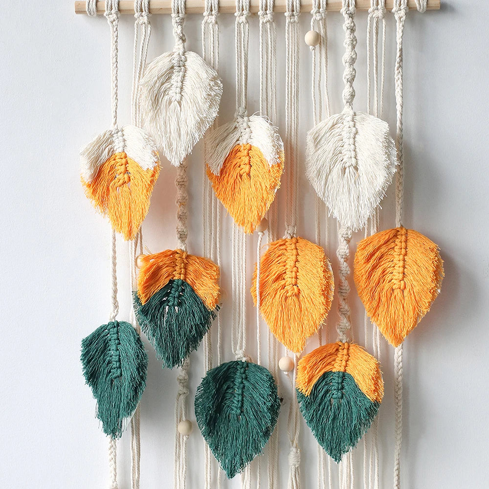 Macrame wall hanging with leaf-shaped decorations in orange, green, and white on a light gray background.