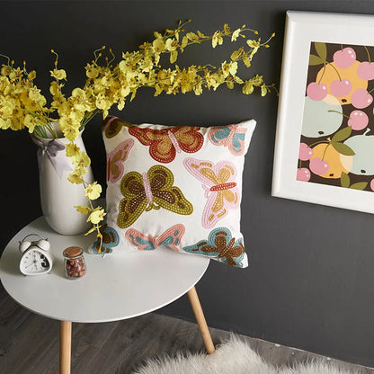 Decorative pillow with butterfly pattern on a small round table against a dark wall with a framed picture. floral embroidery on linen cotton cushion cover – boho home decor accent