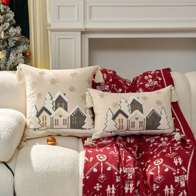 Decorative pillows with house patterns on a couch with a red blanket, Christmas tree in the background.