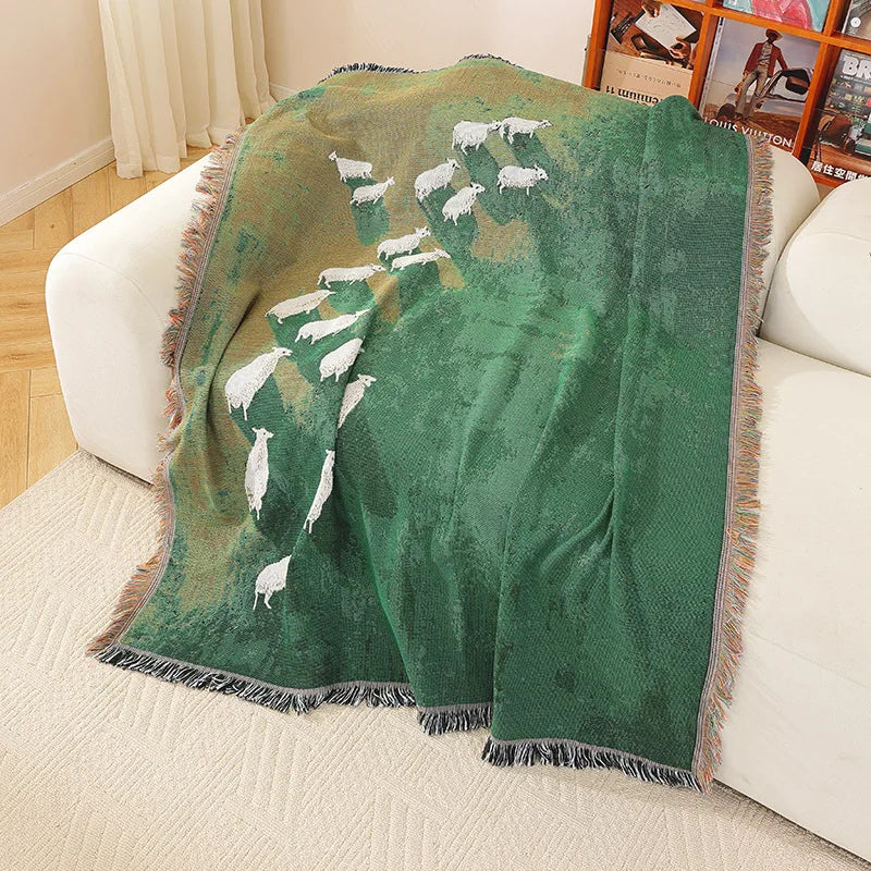 Green blanket with sheep design draped over a couch in a living room. Grassland sheep tassel blanket, woven scenic throw for sofa, bed, or outdoor picnic, boho-inspired home decor blanket.