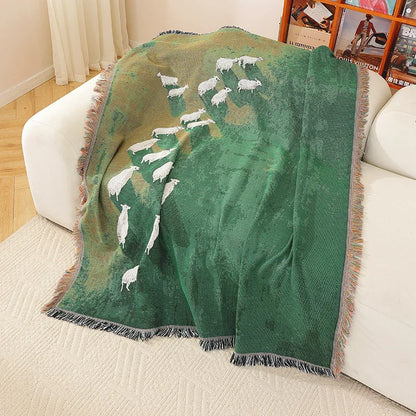 Green blanket with sheep design draped over a couch in a living room. Grassland sheep tassel blanket, woven scenic throw for sofa, bed, or outdoor picnic, boho-inspired home decor blanket.