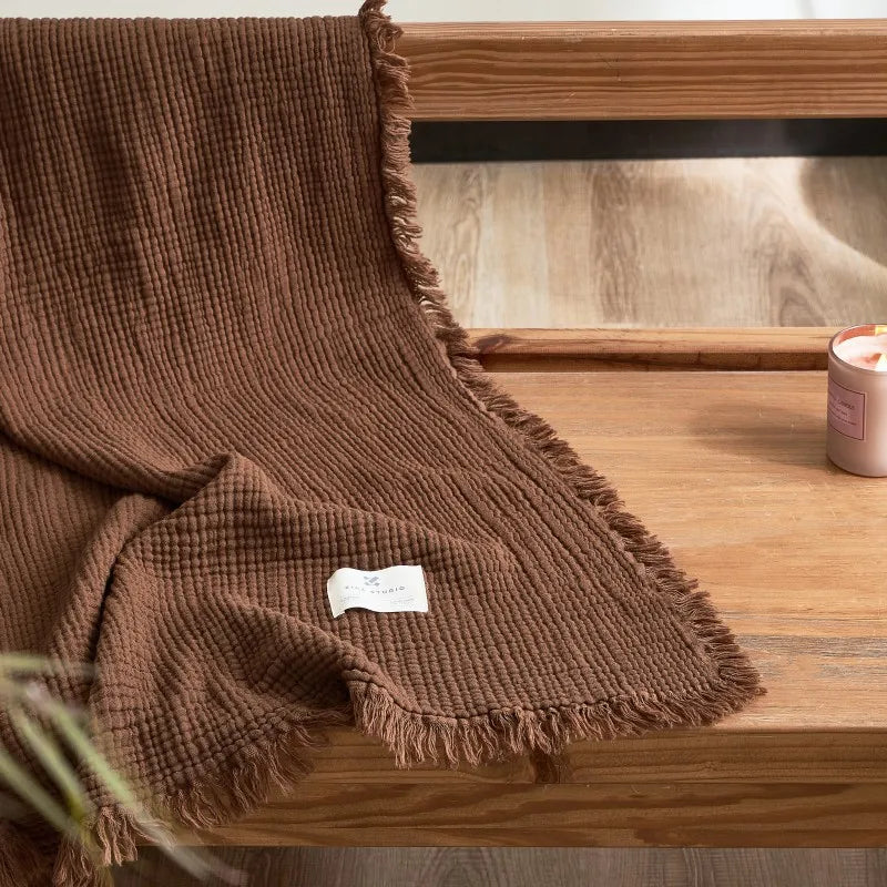 Brown textured blanket draped over a wooden surface with a candle in the background. Maillard cotton gauze blanket with tassels, four-layer muslin throw for sofa, bed, and boho home decor