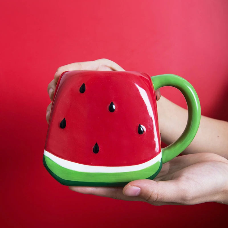 Hand holding a red watermelon-shaped mug with green handle against a red background Creative watermelon ceramic mug, kawaii coffee and milk cup with hand-painted design, 500ml porcelain drinkware for kitchen decor