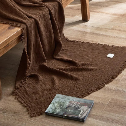 Brown textured blanket draped over a wooden bench on a wooden floor with a book underneath. Maillard cotton gauze blanket with tassels, four-layer muslin throw for sofa, bed, and boho home decor