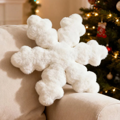 A fluffy white snowflake-shaped throw pillow resting on a beige armchair, with a softly blurred Christmas tree in the background.