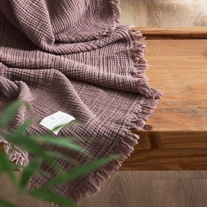 Brown textured blanket on a wooden surface with a blurred background Maillard cotton gauze blanket with tassels, four-layer muslin throw for sofa, bed, and boho home decor