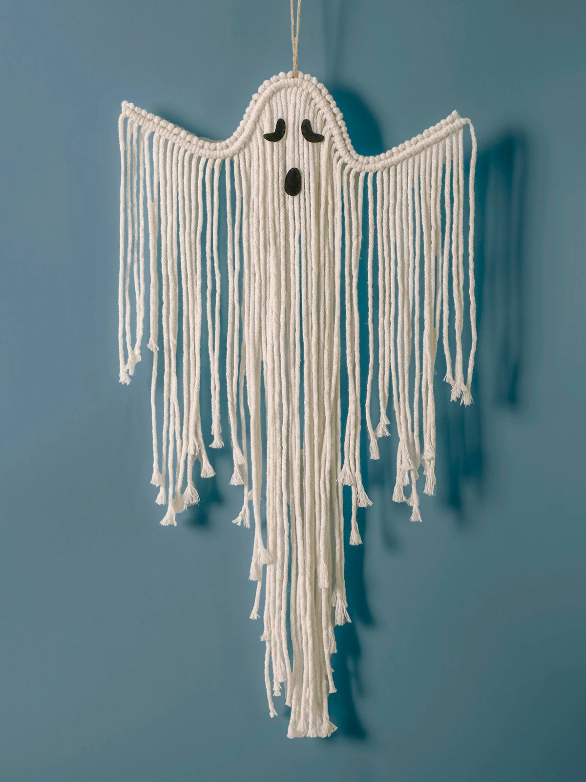 A white macrame ghost wall hanging with long fringe and small black embroidered eyes and mouth, suspended against a solid blue wall from My Boho Home