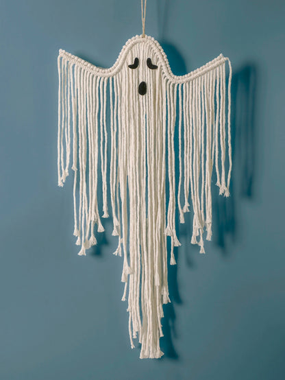 A white macrame ghost wall hanging with long fringe and small black embroidered eyes and mouth, suspended against a solid blue wall from My Boho Home