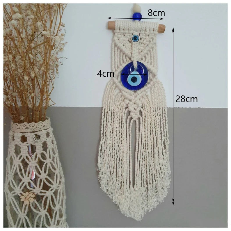 A cream macrame wall hanging with blue glass evil eye beads and wooden dowel, shown with dimension labels 8cm, 4cm, and 28cm. A vase of dried flowers sits to the left against a two-tone white and gray background.