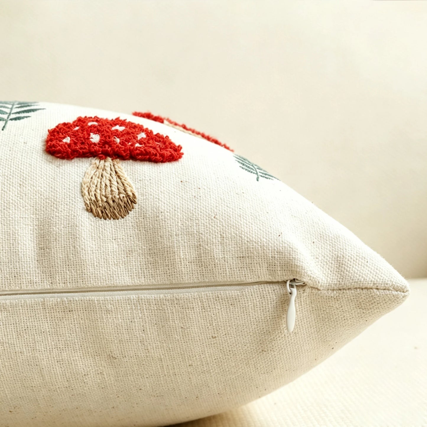 A close-up of a square beige woven throw pillow featuring a red and beige embroidered mushroom design on the front, with a visible zipper closure. Boho fall home decor.