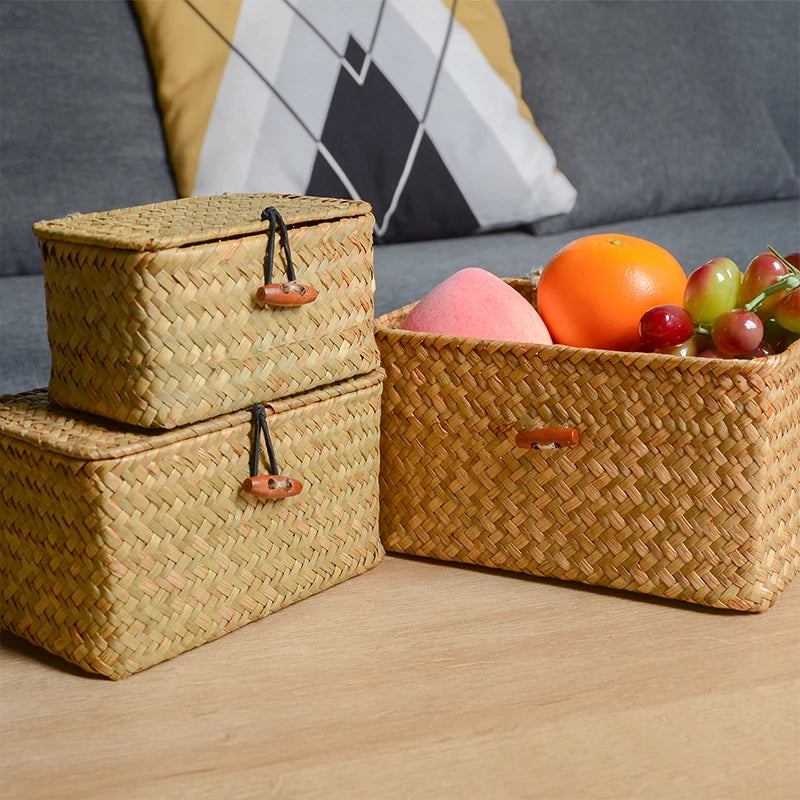 Woven storage baskets with a lid on a wooden surface, one containing fruits. Handmade seagrass rattan storage box with lid, rectangular woven organizer basket for makeup, jewelry, office, and boho home decor