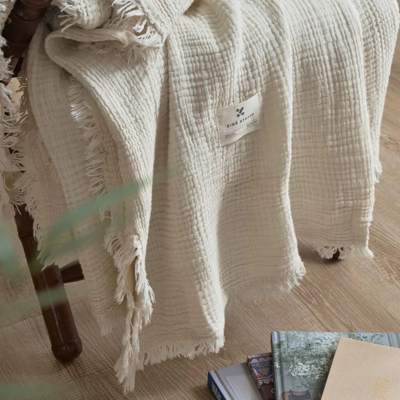 Beige textured blanket draped over a chair with a visible brand label on a wooden floor. Maillard cotton gauze blanket with tassels, four-layer muslin throw for sofa, bed, and boho home decor