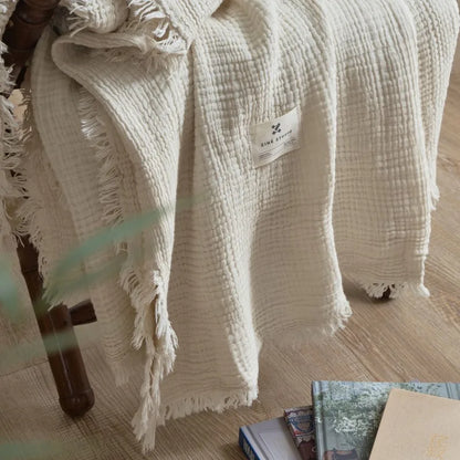Beige textured blanket draped over a chair with a visible brand label on a wooden floor. Maillard cotton gauze blanket with tassels, four-layer muslin throw for sofa, bed, and boho home decor