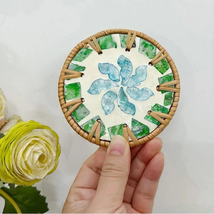 Hand holding a round woven coaster with green and blue leaf patterns on a light gray background