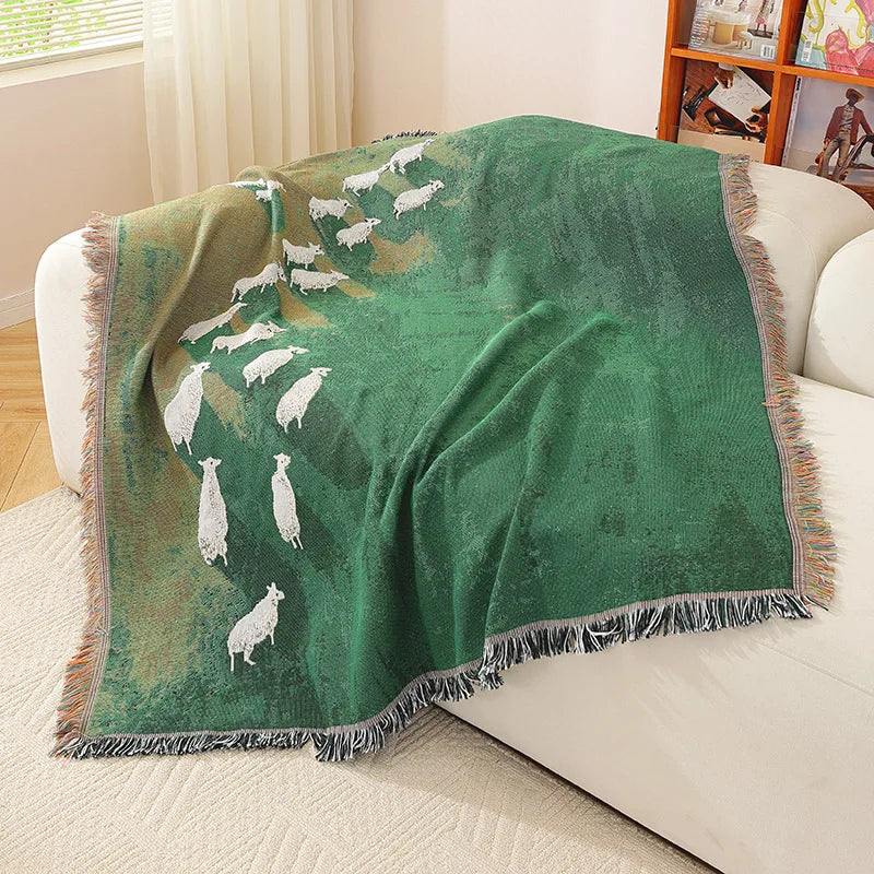 Green blanket with white animal pattern draped over a white couch in a living room. Grassland sheep tassel blanket, woven scenic throw for sofa, bed, or outdoor picnic, boho-inspired home decor blanket.